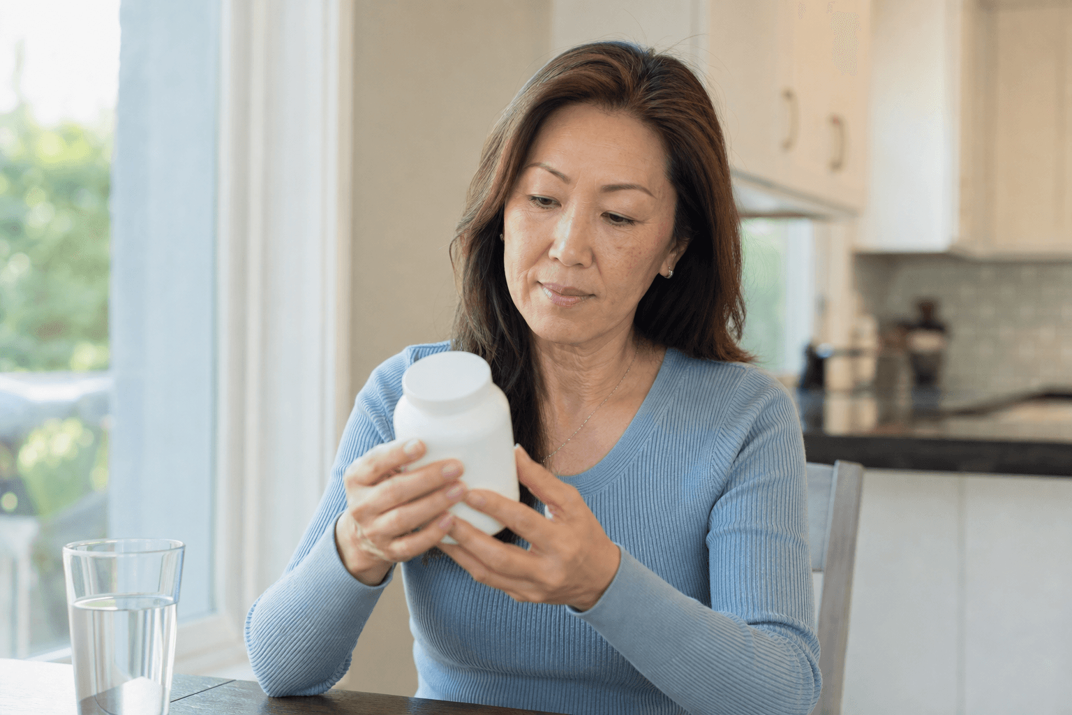 Mature woman reading an unbranded supplement bottle at a kitchen table